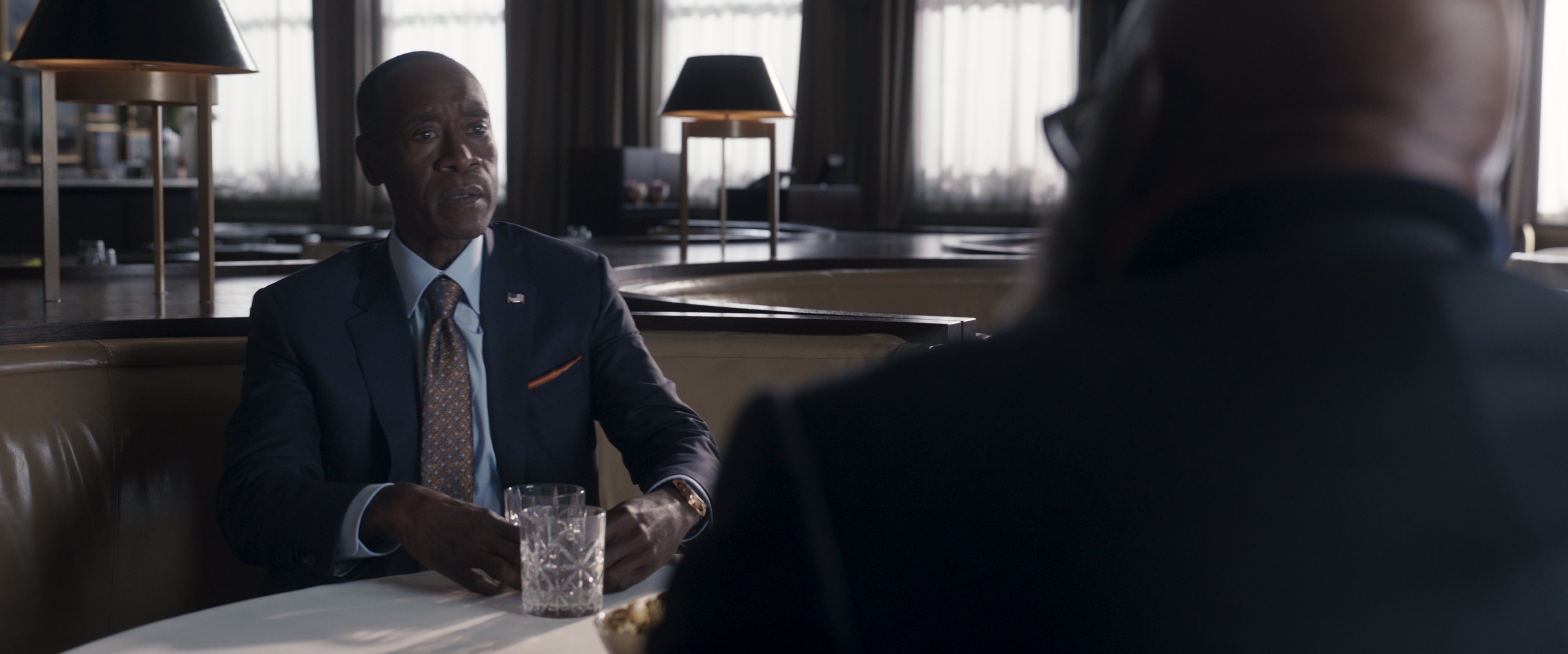 Still image of Rhodey wearing a suit and sitting at a white tableclothed table facing Nick Fury