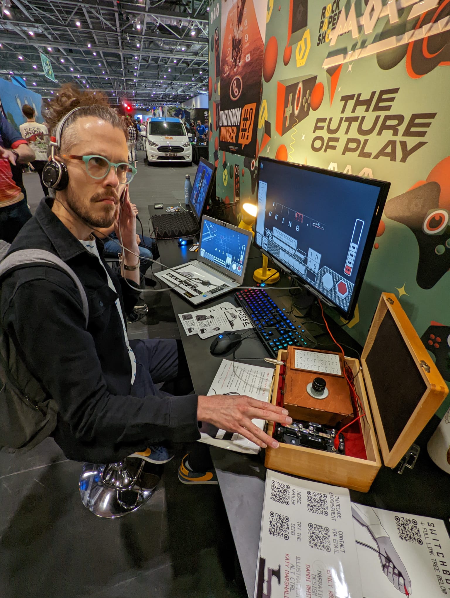 Bertie playing a Morse Code game at EGX. There's a wooden box on a bench with the familiar little Morse Code clicker on it, and a big launcher button nearby. Bertie looks rapt.