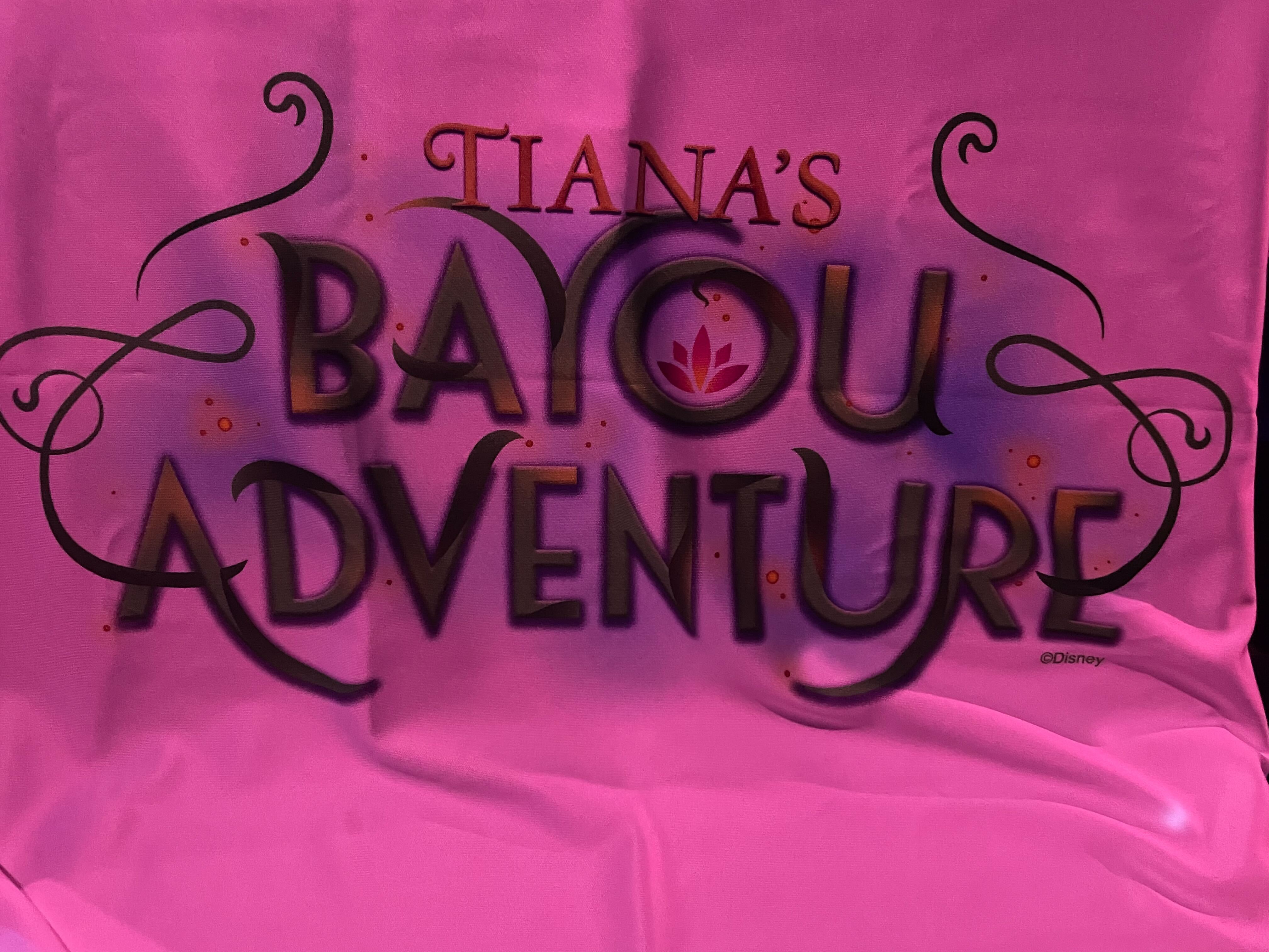 Pink tinted photograph of a piece of fabric that reads Tiana's Bayou Adventure