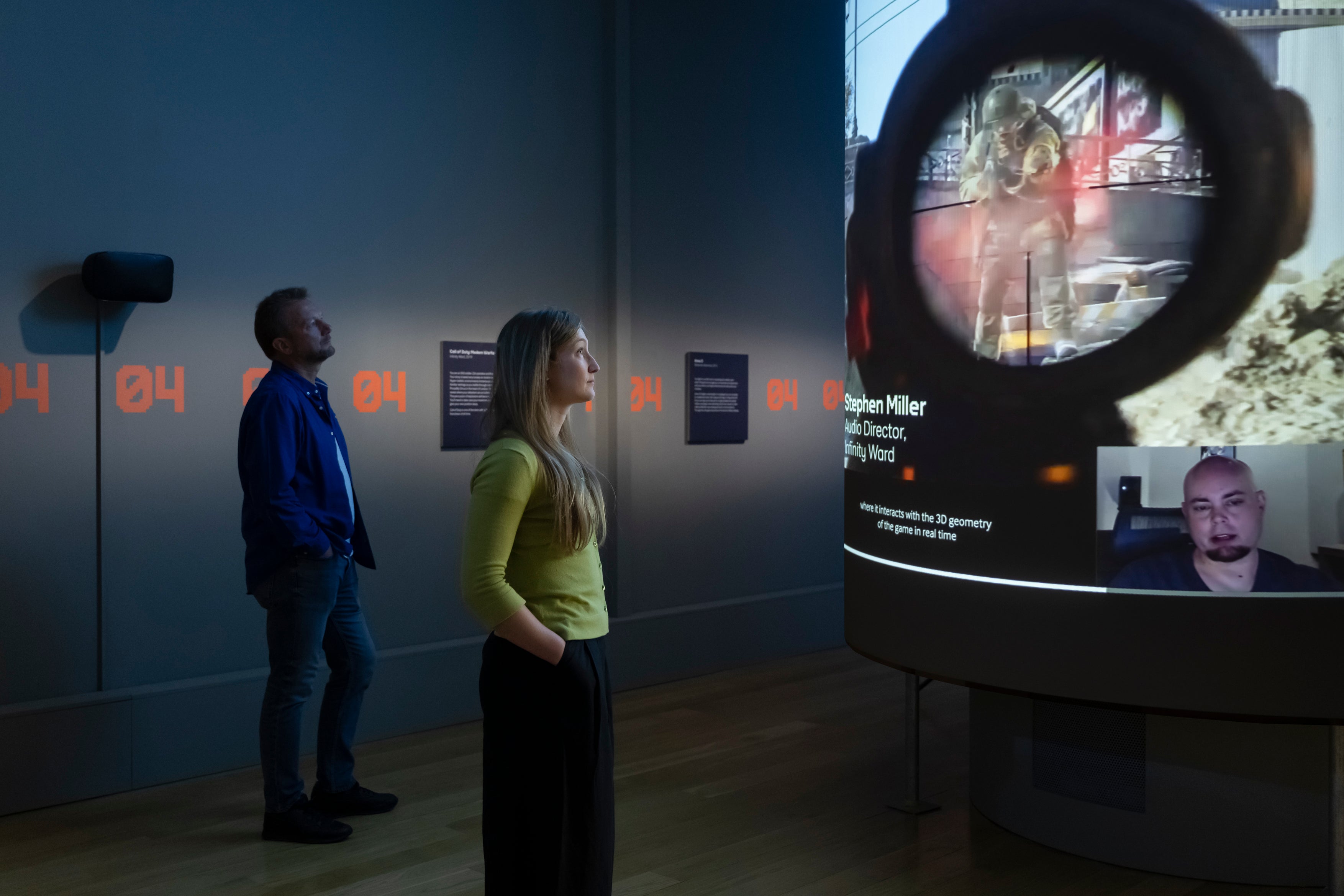 Museum visitors stare at a screenshot that shows a video game soldier bleeding in the crosshairs of a sniper scope.
