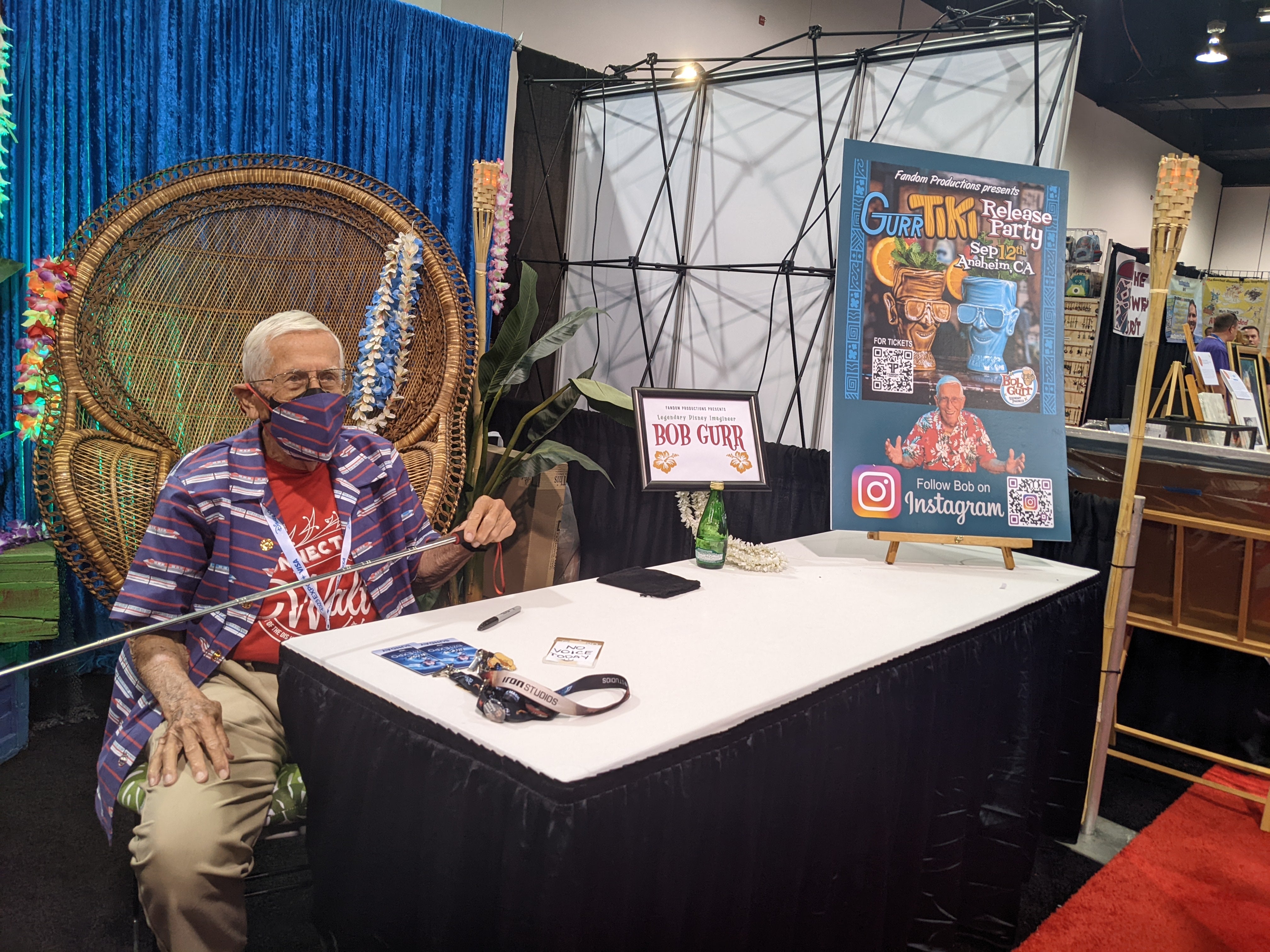 Bob Gurr wearing a mask, sitting in a large wicker chair and holding a telescoping stick to his right