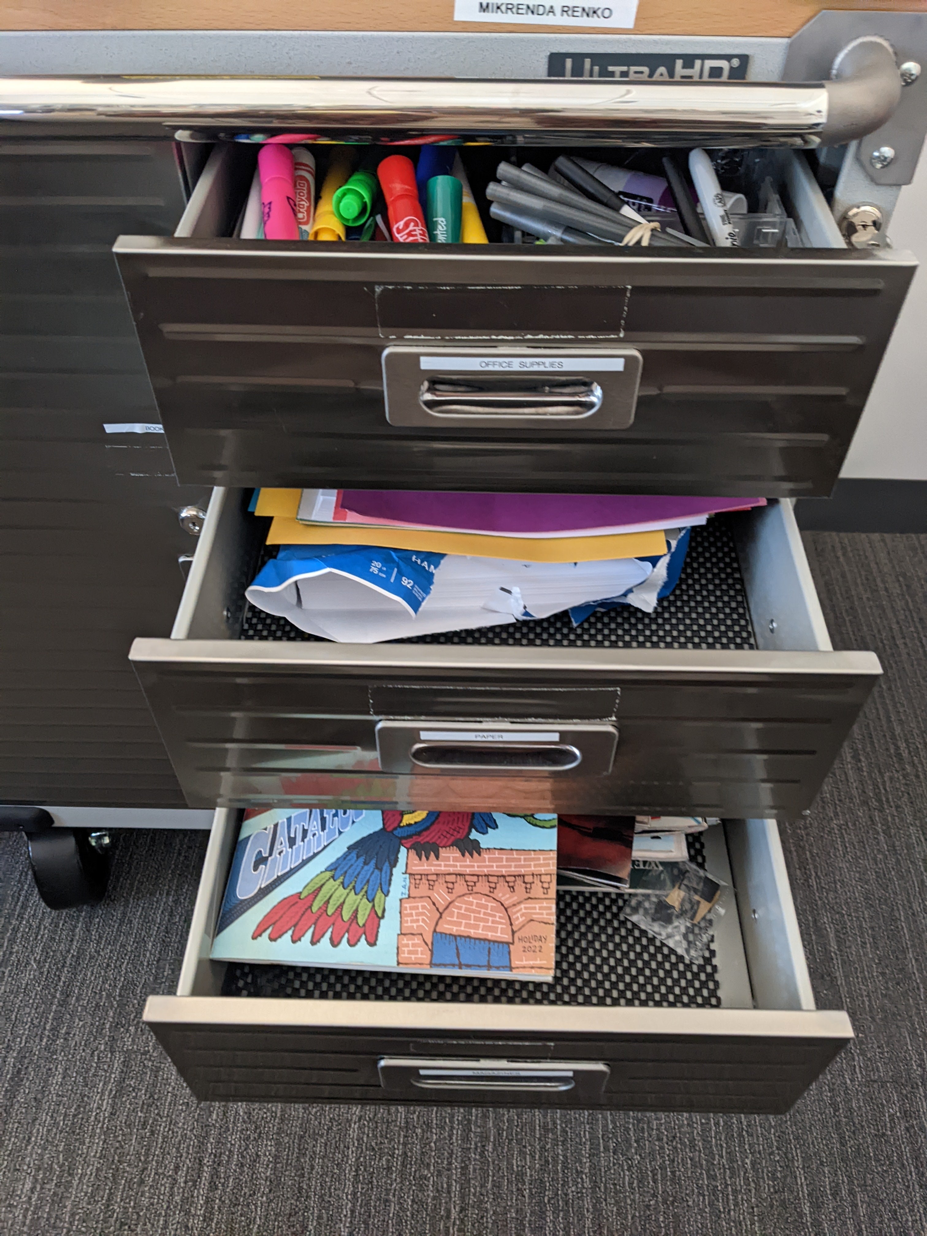 A photograph of a gunmetal grey cart with three open drawers featuring magazines, collaging materials, markers