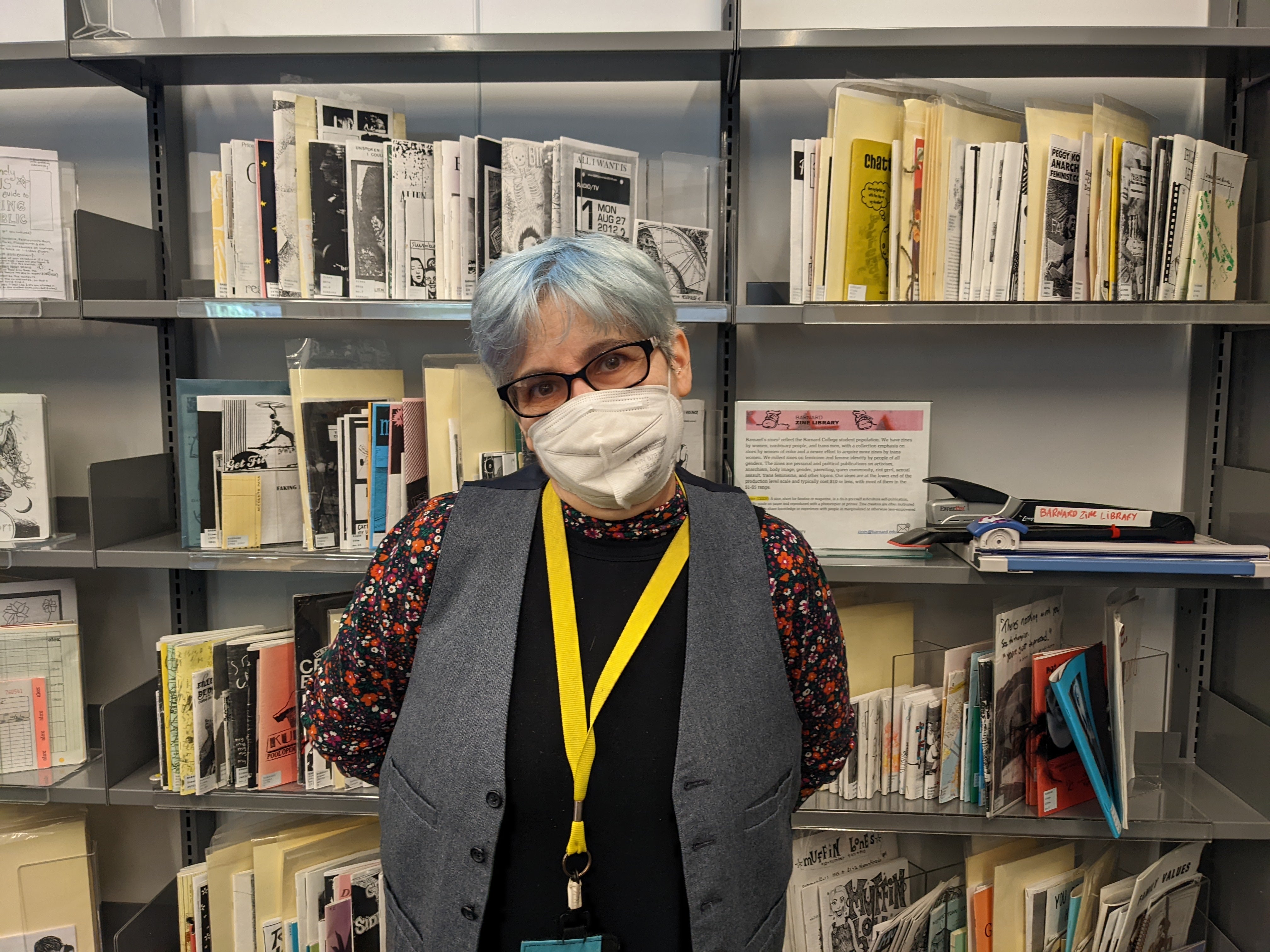 Zine librarian Jenna Freedman with short colored hair, wearing glasses, a mask, and a yellow lanyard in front of shelves featuring zines