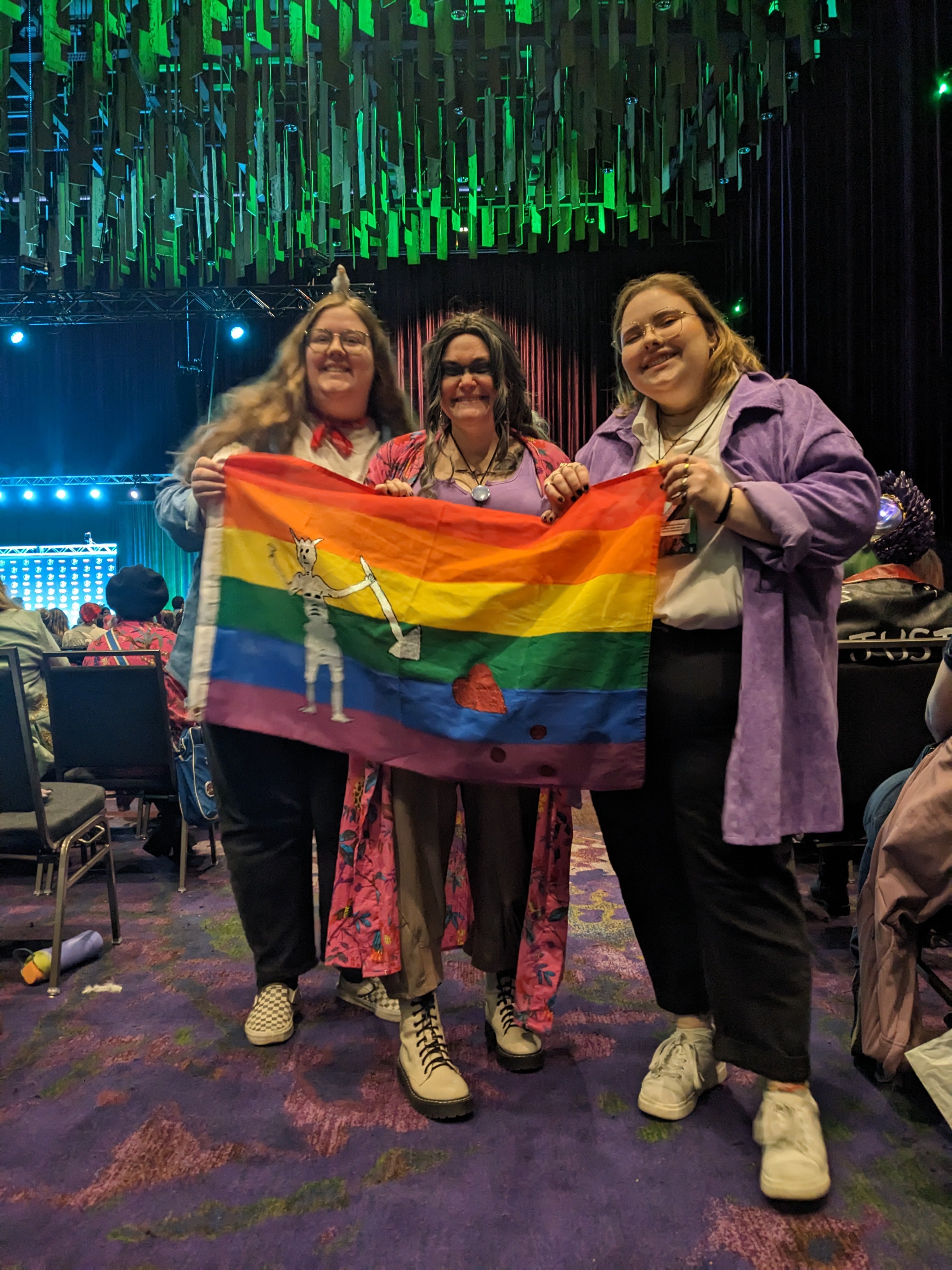 Three people in cosplay holding a rainbow flag
