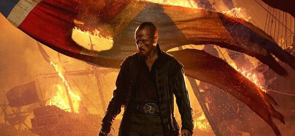 Toby Stephens as Captain Flint in front of a burning background and burning British flag