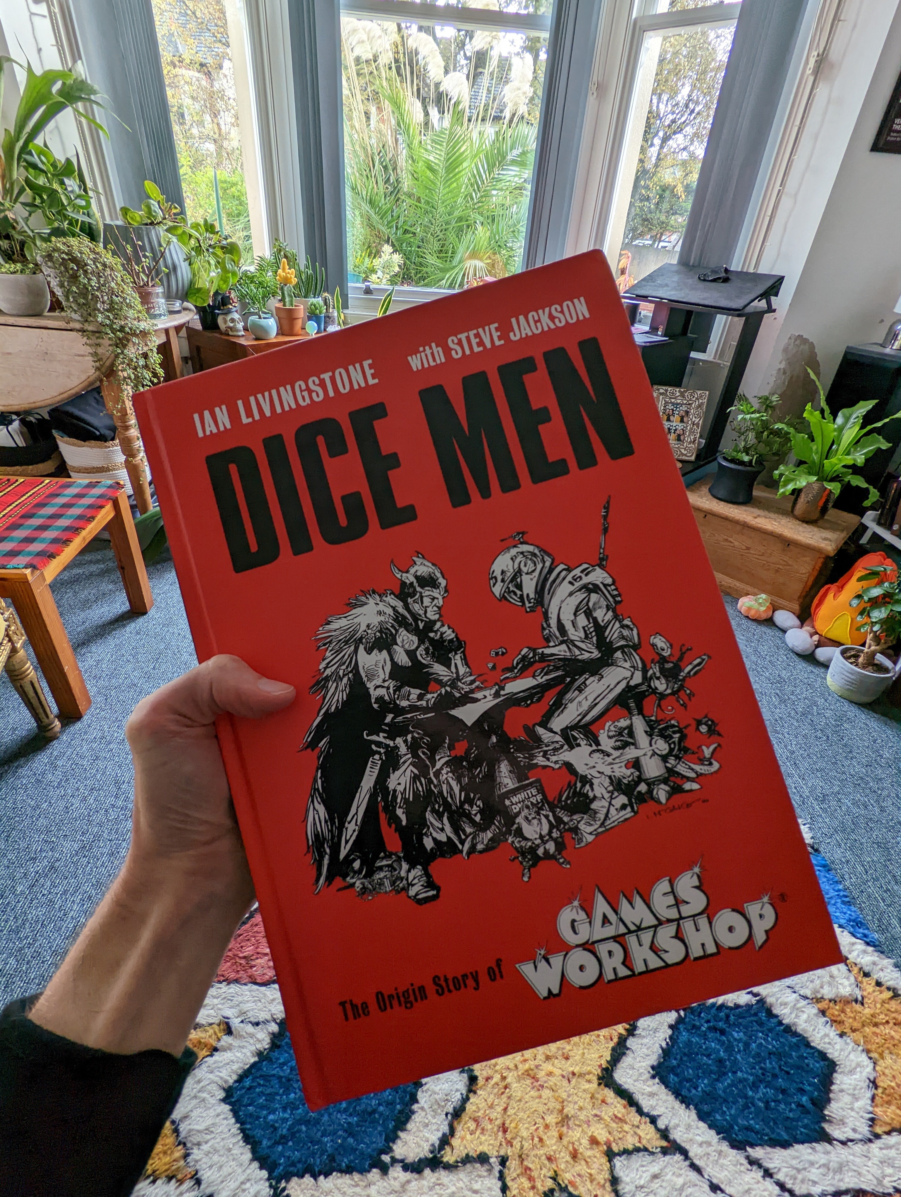 Bertie's hand, and adjoining arm, holding up the Dice Men Games Workshop history book. It's bright red with two characters - one from fantasy, one from sci-fi - playing a game of dice together. Bertie's  flat can be pictured in the background. It has some plants in it.