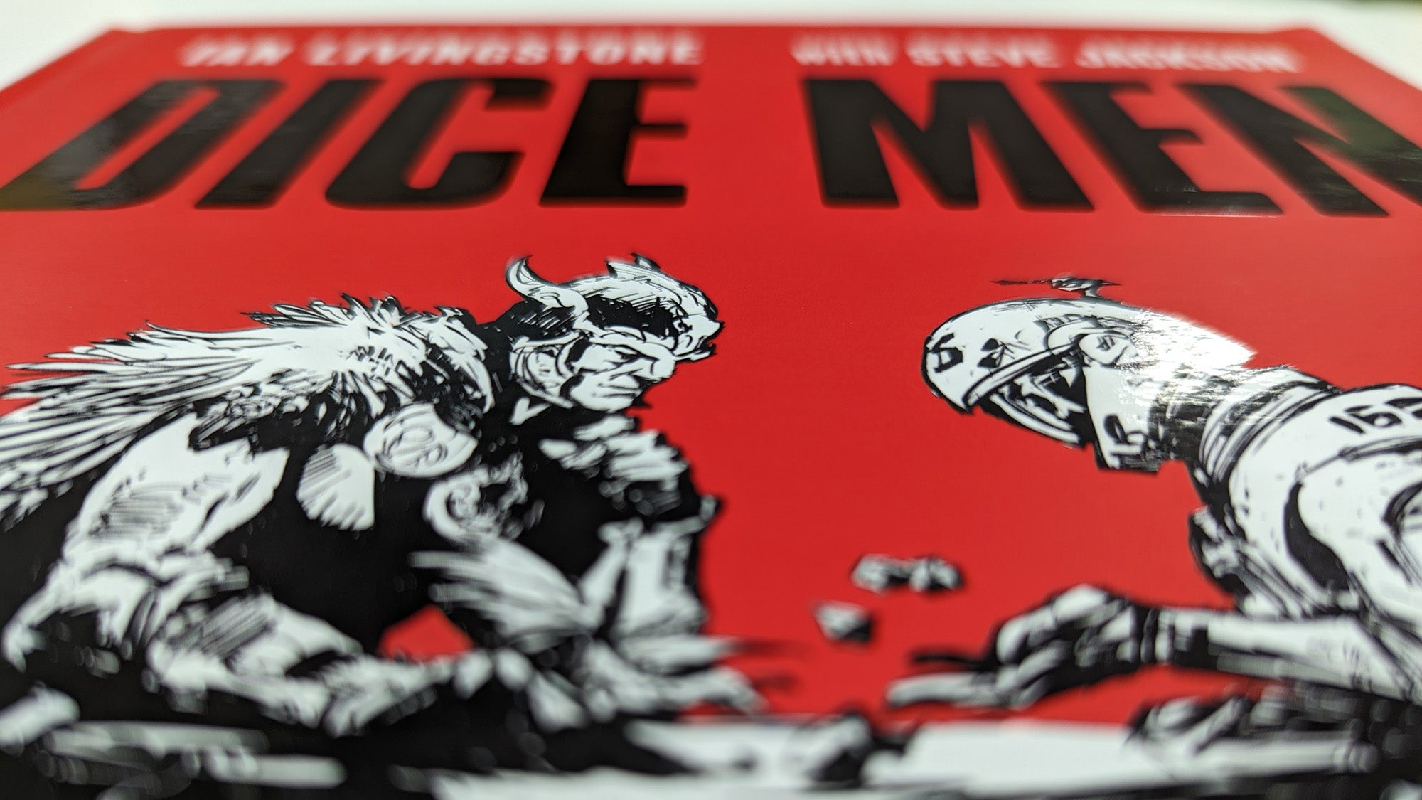 A photo of the Dice Men Games Workshop history book. It's red with two characters fantastical characters playing dice on the front cover. It's the classic Ian McCaig illustration that was on early Games Workshop bags.
