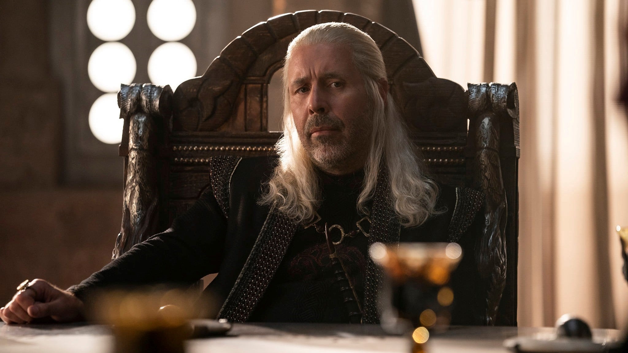 Paddy Considine as King Viserys in HBO television series House of the Dragon. He sits in his council chamber chair, hands resting on the table in front of him.