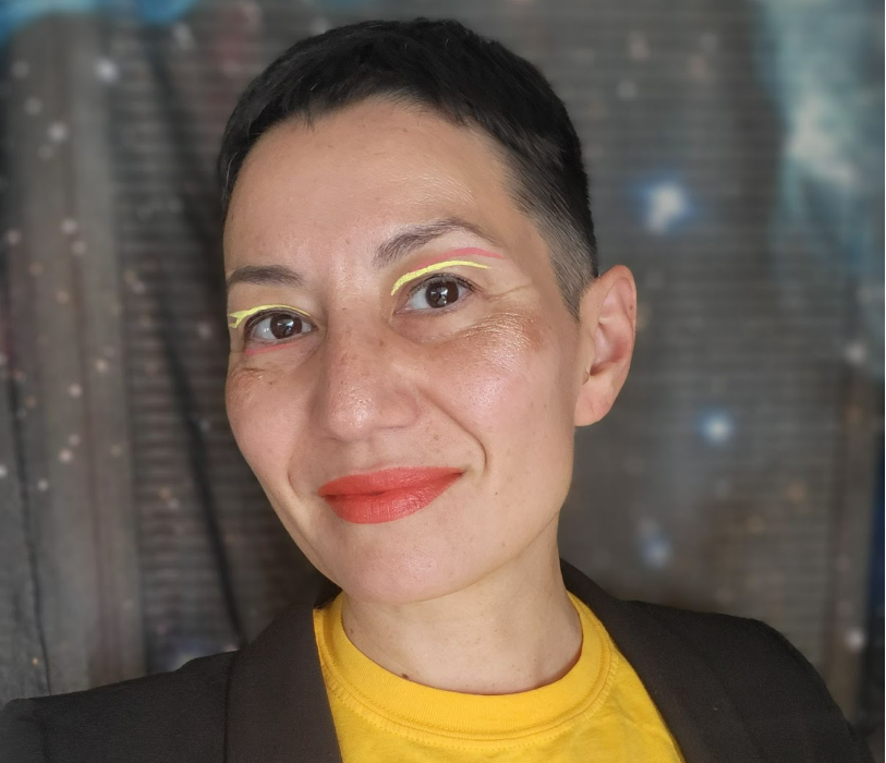Profile photo of MariNaomi, a queer Japanese American person with light skin and shorn black hair, wearing a dark blazer over a yellow t-shirt that says "Asian Love" in red-orange. They are posed in front of a dark and starry outer space backdrop.