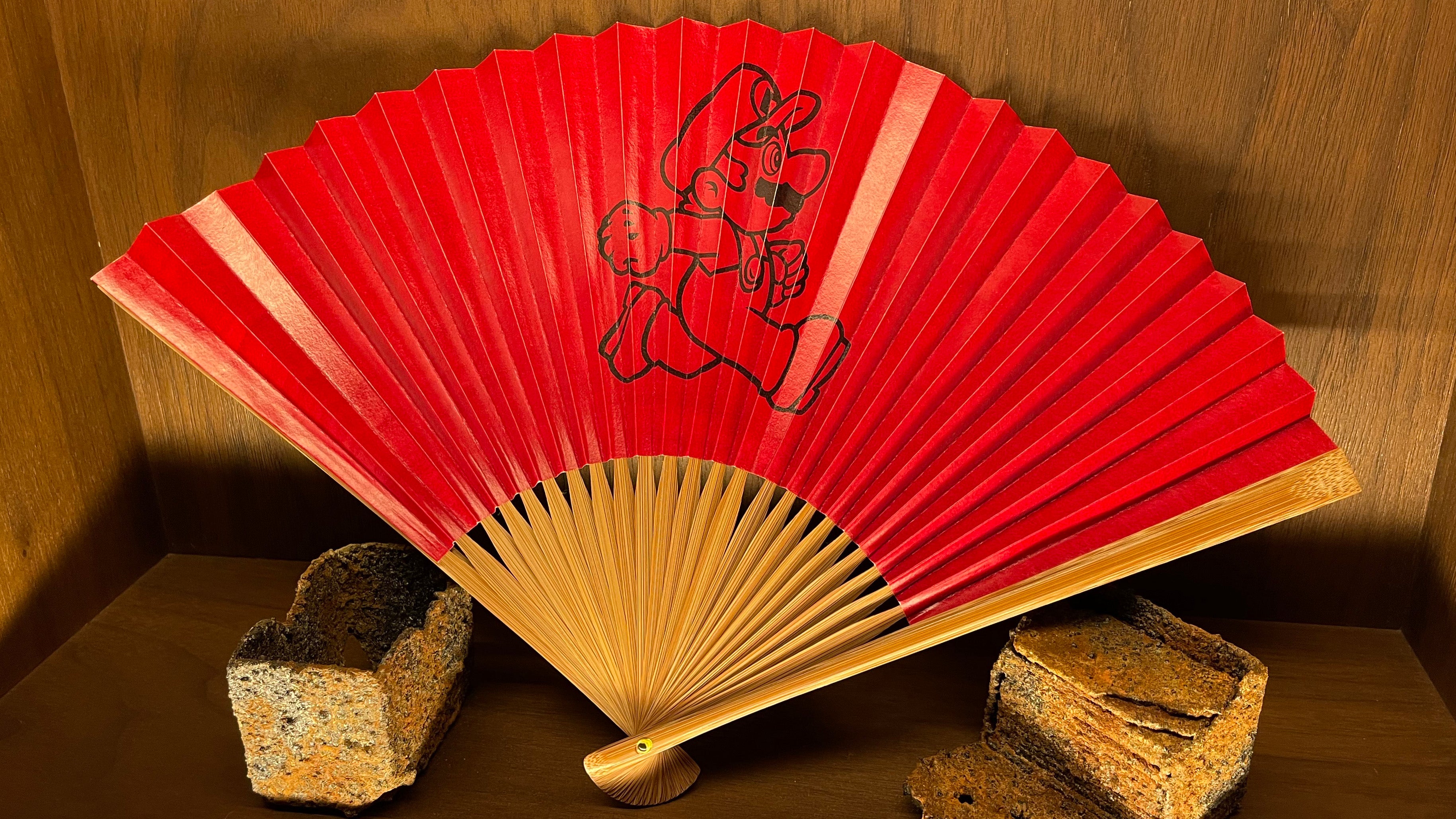 A wooden fan spread open and occupying a bookshelf. Two-thirds of the fan is coloured red, and in the middle of the fan-spread is a picture of Mario, in a running posture.