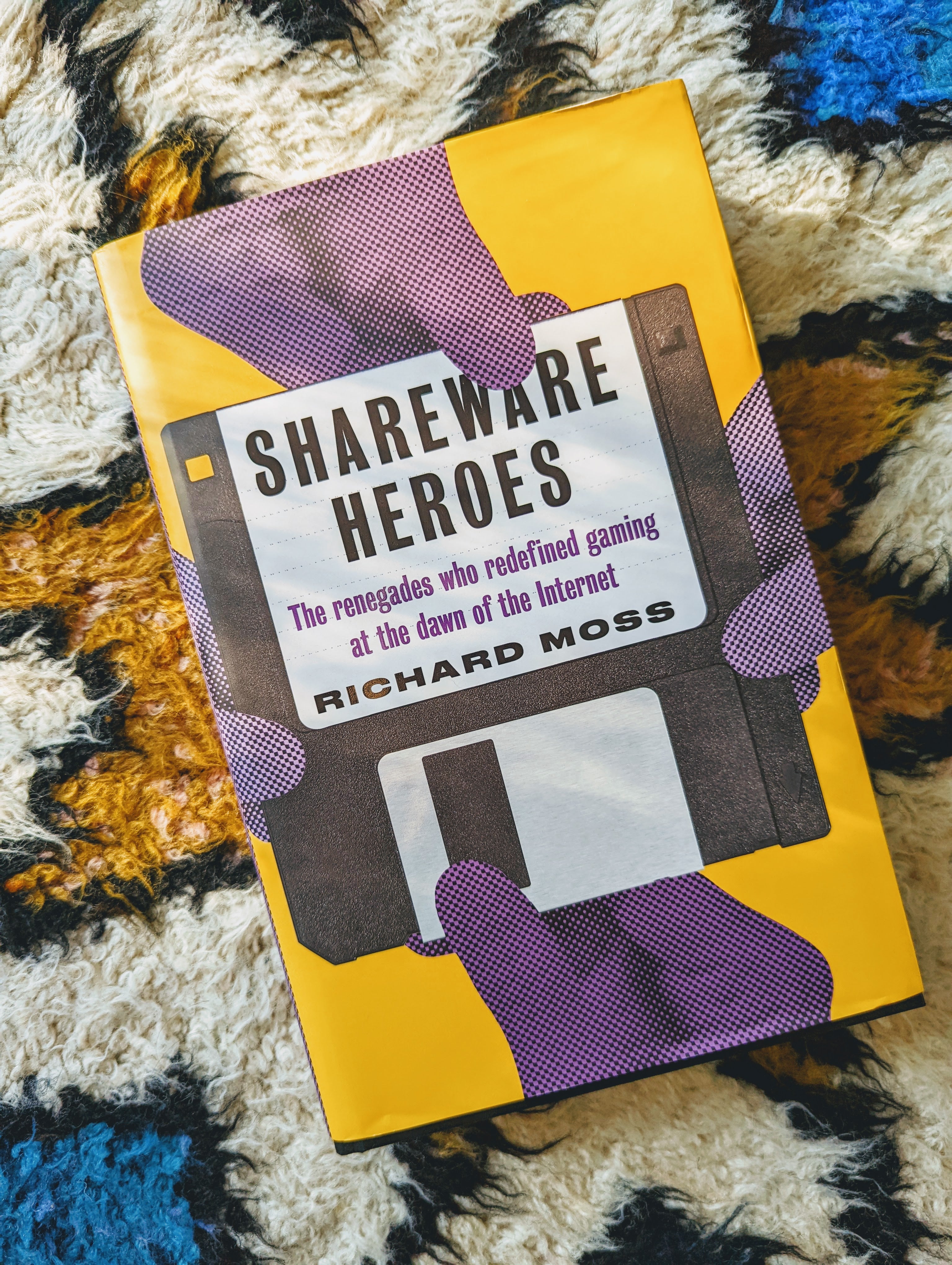 Bertie'nin harika çok renkli halısında resmedilen Shareware Heroes kitabı.  Kitap kapağında, kitabın ve yazar Richard Ross'un adını taşıyan bir disketi çeken dört mor el vardır.  Kitabın geri kalanı sarı renklidir.