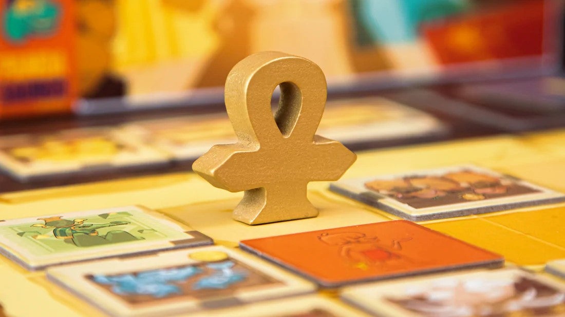 A close-up picture of a golden-coloured, wooden ankh, on a sandy-coloured game board with some tiles placed face-up around it.