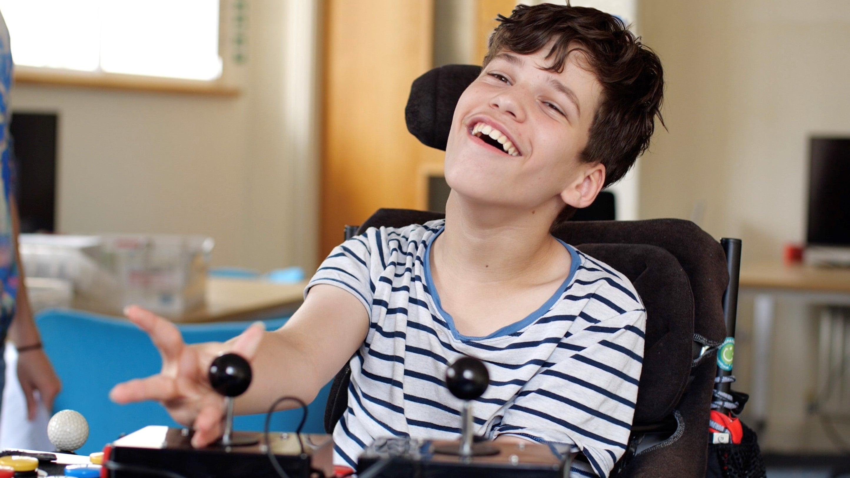 A boy with physical disabilities using bespoke gaming controllers to play a game. He is delighted and his smile says a thousand words.