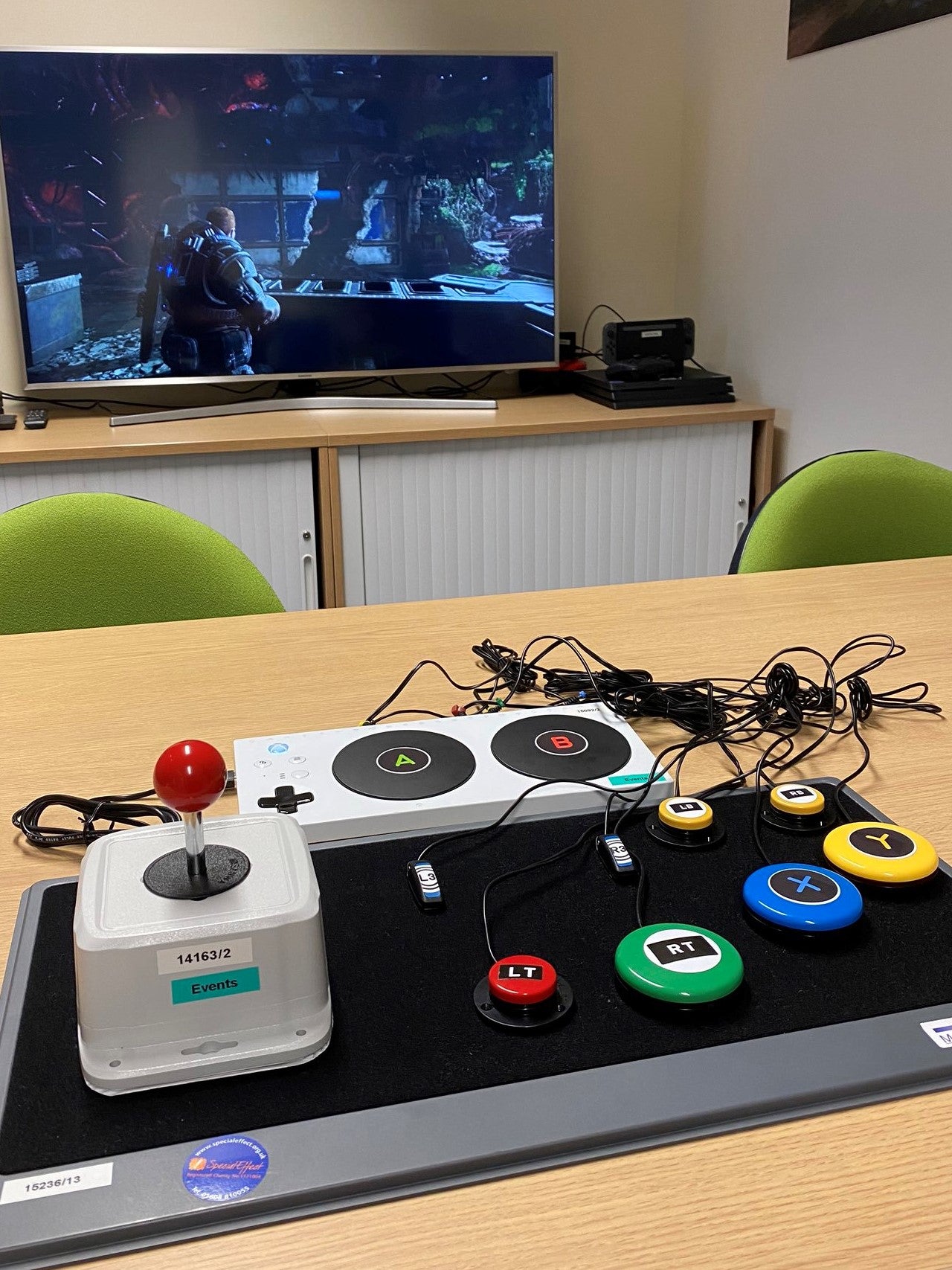 A close-up of a bespoke SpecialEffect controller set-up on a table. There's a joystick, there are several big buttons scattered nearby, and there's the Xbox Adaptive Controller, which everything is wired into. In the background, Gears of War is playin gon a TV.