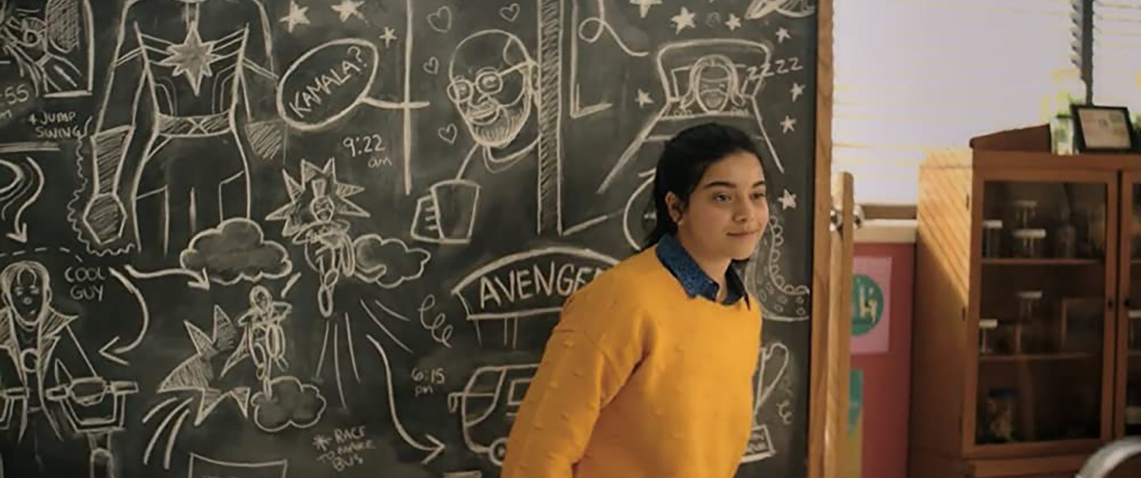 Ms. Marvel still Iman Vellani as Kamala Khan in front of a blackboard filled with superhero based doodles