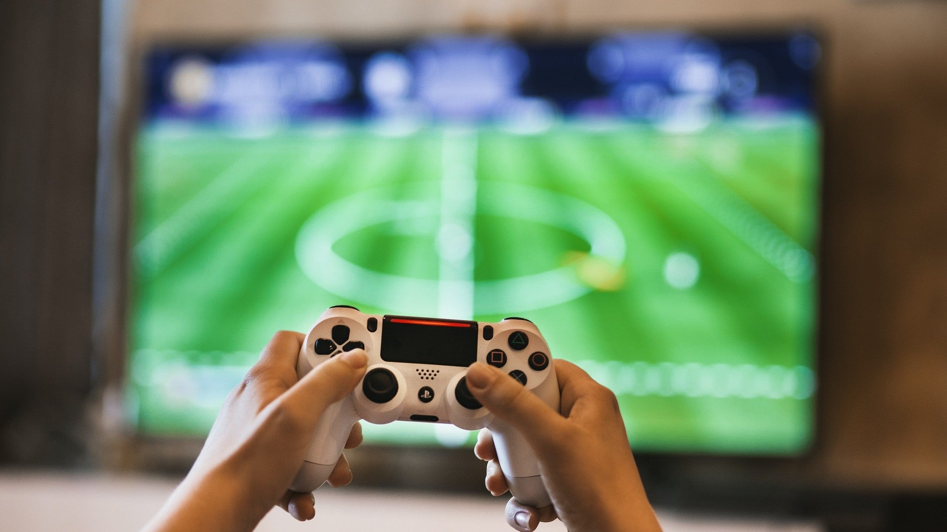 Hands holding a PS4 controller in the foreground, an out of focus screen with a soccer game on it in the background