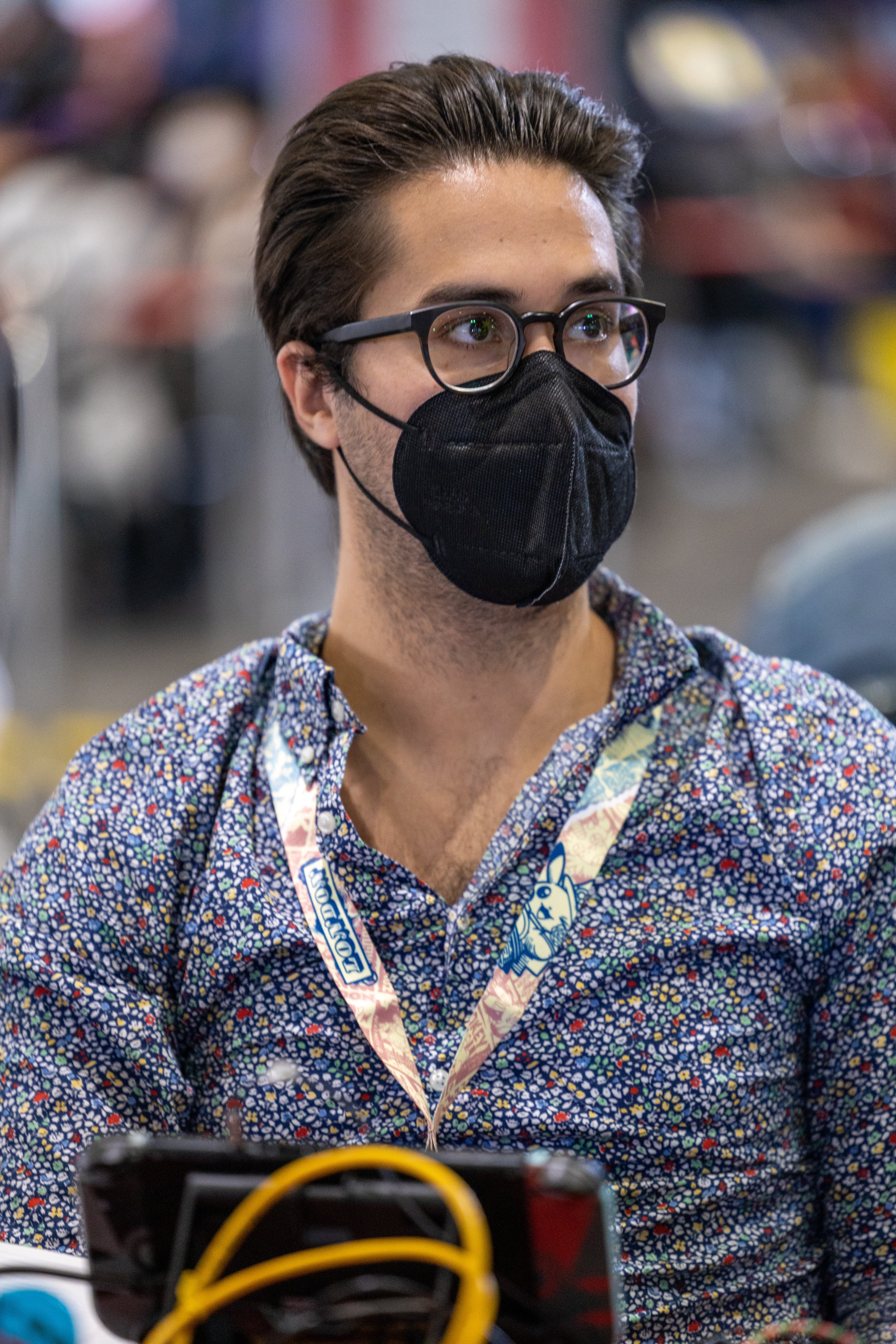 A portrait photograph of Pokemon champion Wolfe Glick looking off to the camera's right. He's nicely groomed, with hair brushed back and a patterned shirt on. A black face mask and glasses cover much of his face.